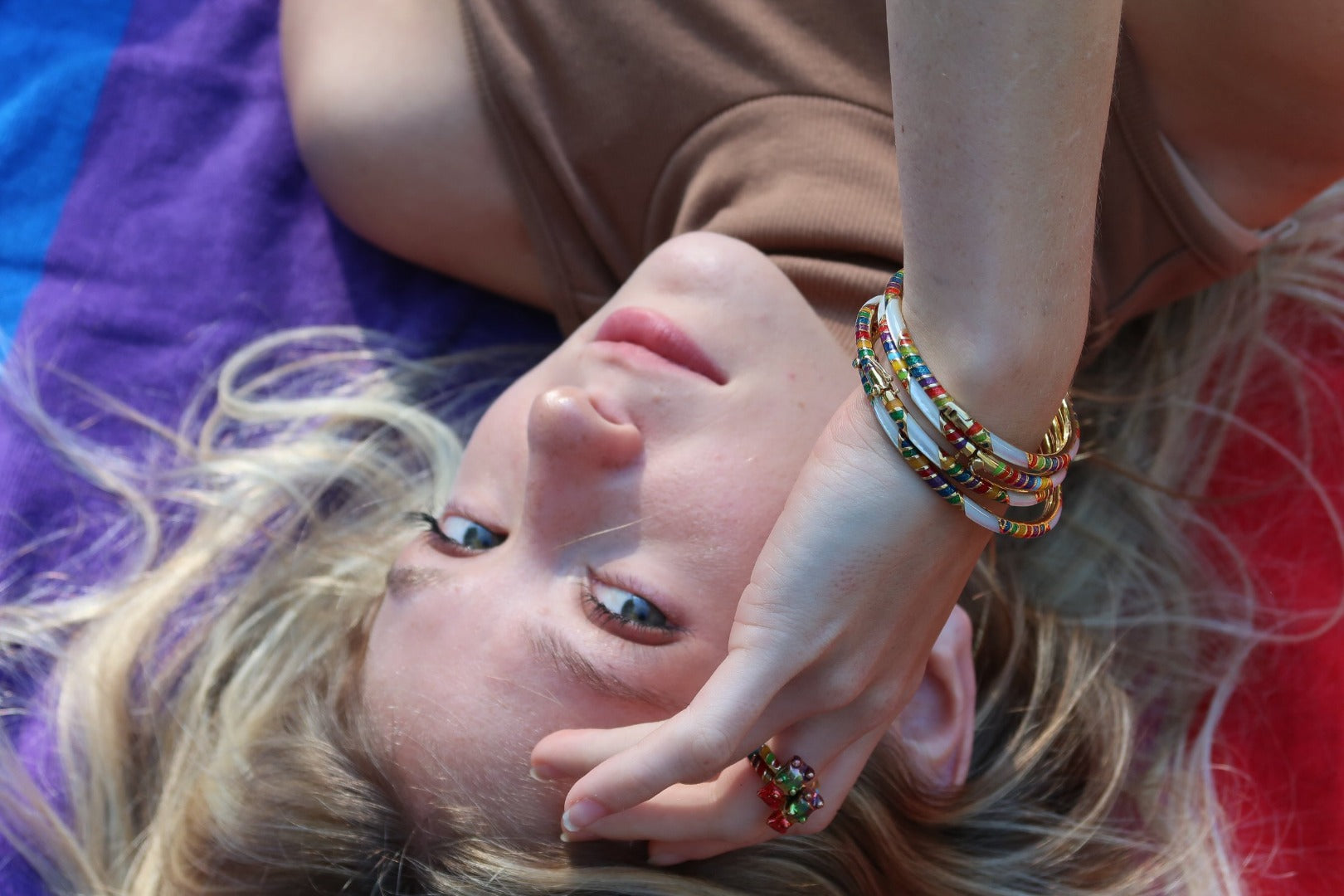 Female model laying on a striped rainbow towel and looking up at the camera with her placed on her forehead. Model is wearing Jan Leslie's SINGLE Rainbow Gummy Bear Sterling Silver Ring with 18K Gold Vermeil and three Stripe Gemstone & Enamel Sterling Silver Stackable Bangle Bracelets in rainbow on gold. 